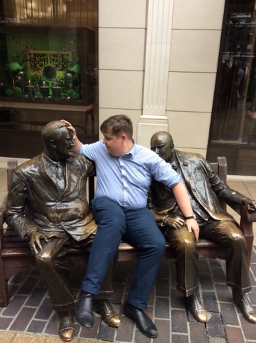 An image of a man taking a picture with 2 statues on a bench